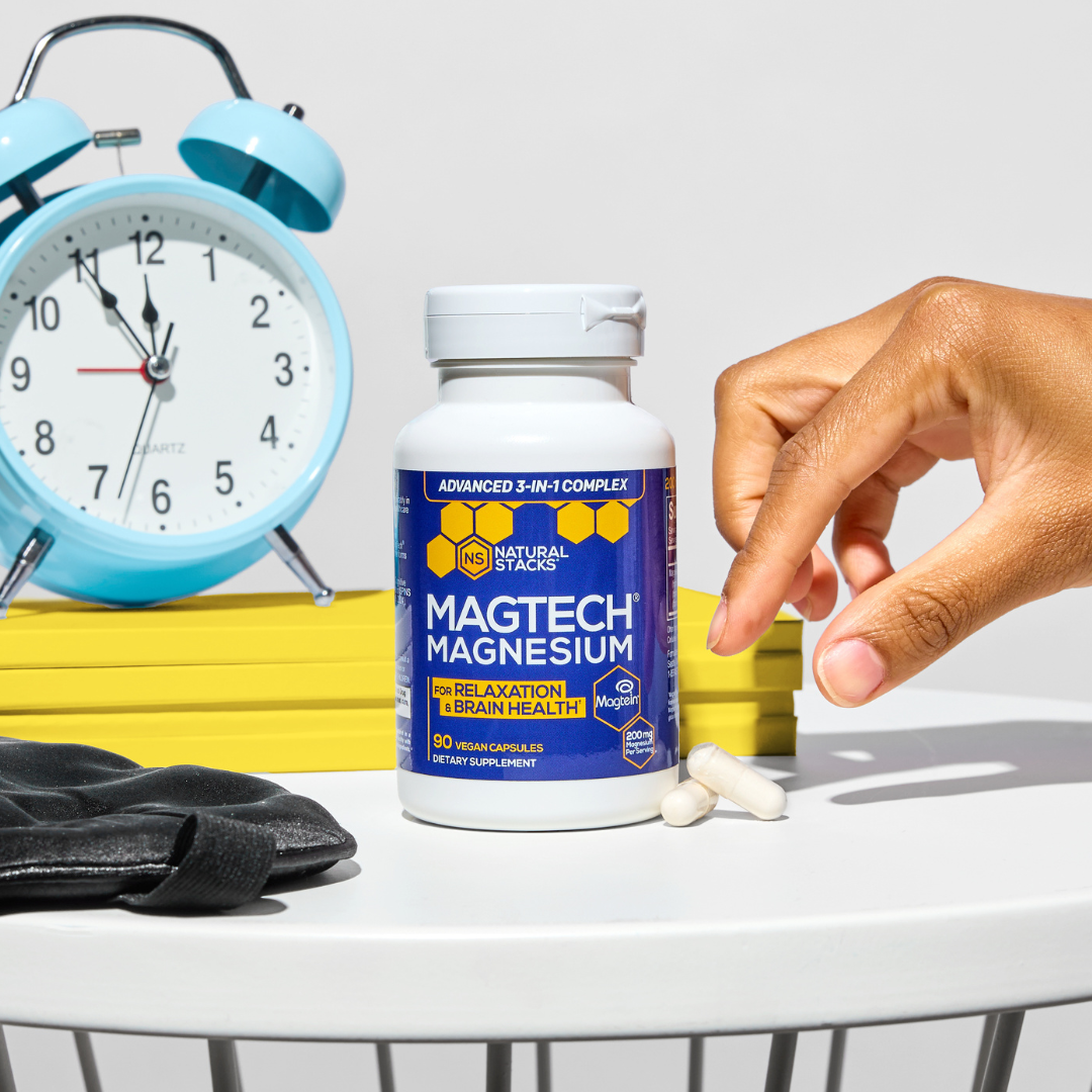 Hand reaching for capsules beside magnesium supplement bottle and blue alarm clock.