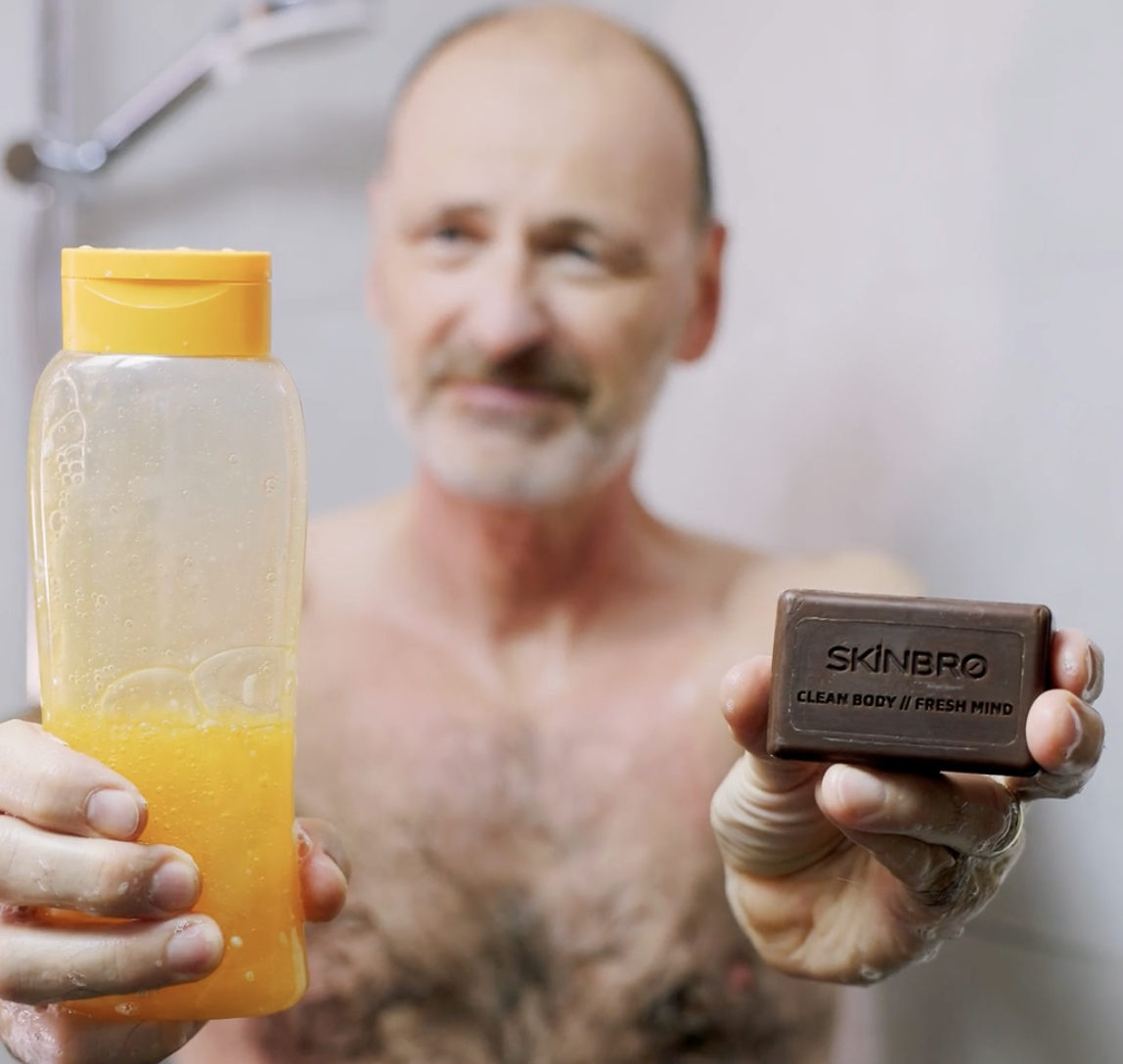 Man holding a bottle of liquid soap and a SKINBRO soap bar in a shower.