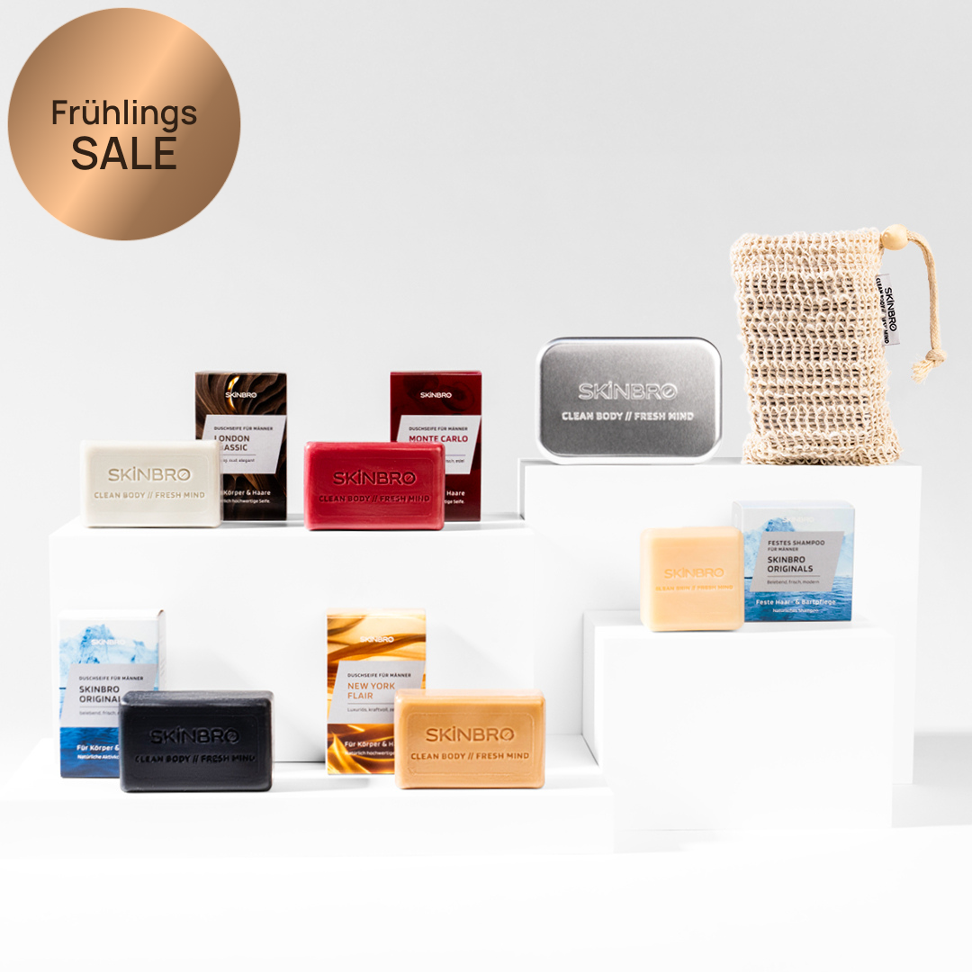 Various colorful SKINBRO soap bars, packaging, a metal tin, and a mesh bag displayed on white blocks.