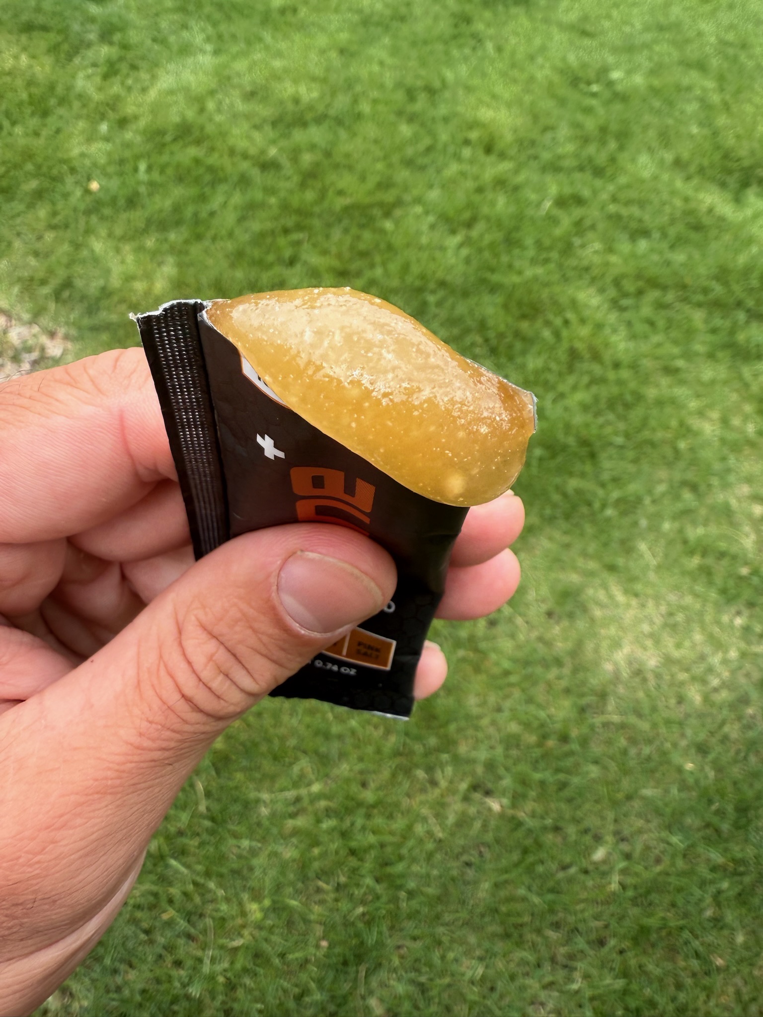A hand holds an open packet, squeezing out a golden-colored energy gel over a grassy background.