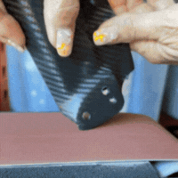 Hands sanding a black object on a belt sander.