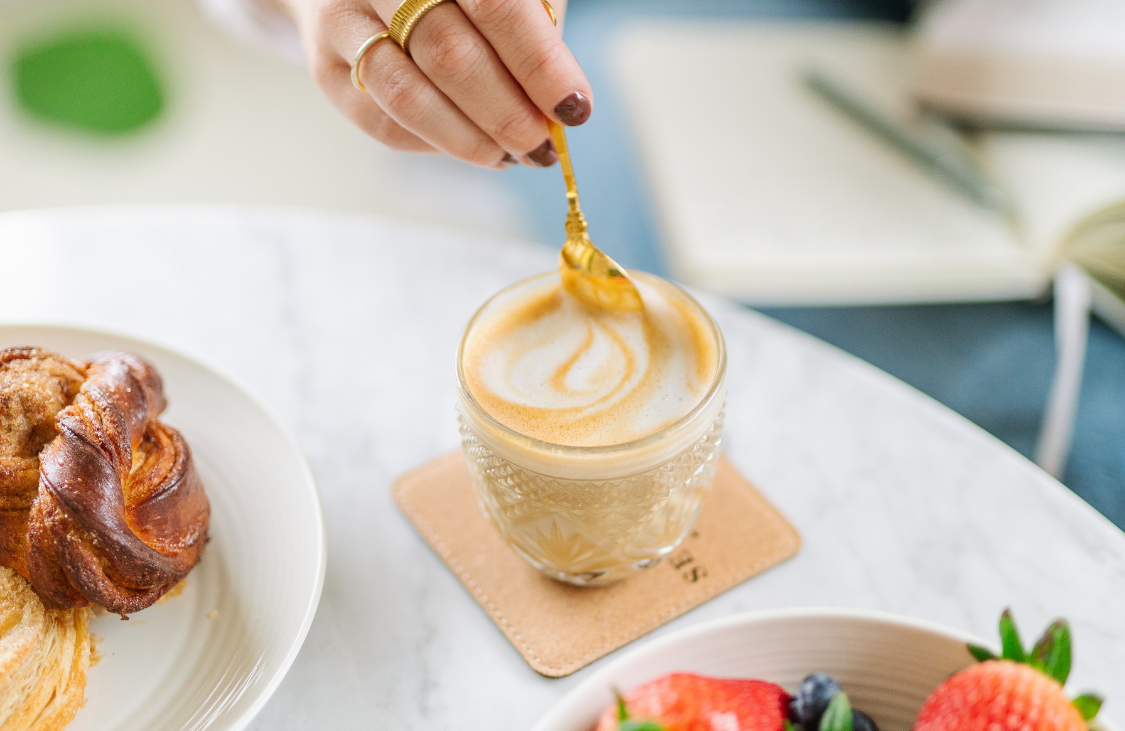 A person's hand wearing gold rings stirs a latte in a decorative glass with a golden spoon.