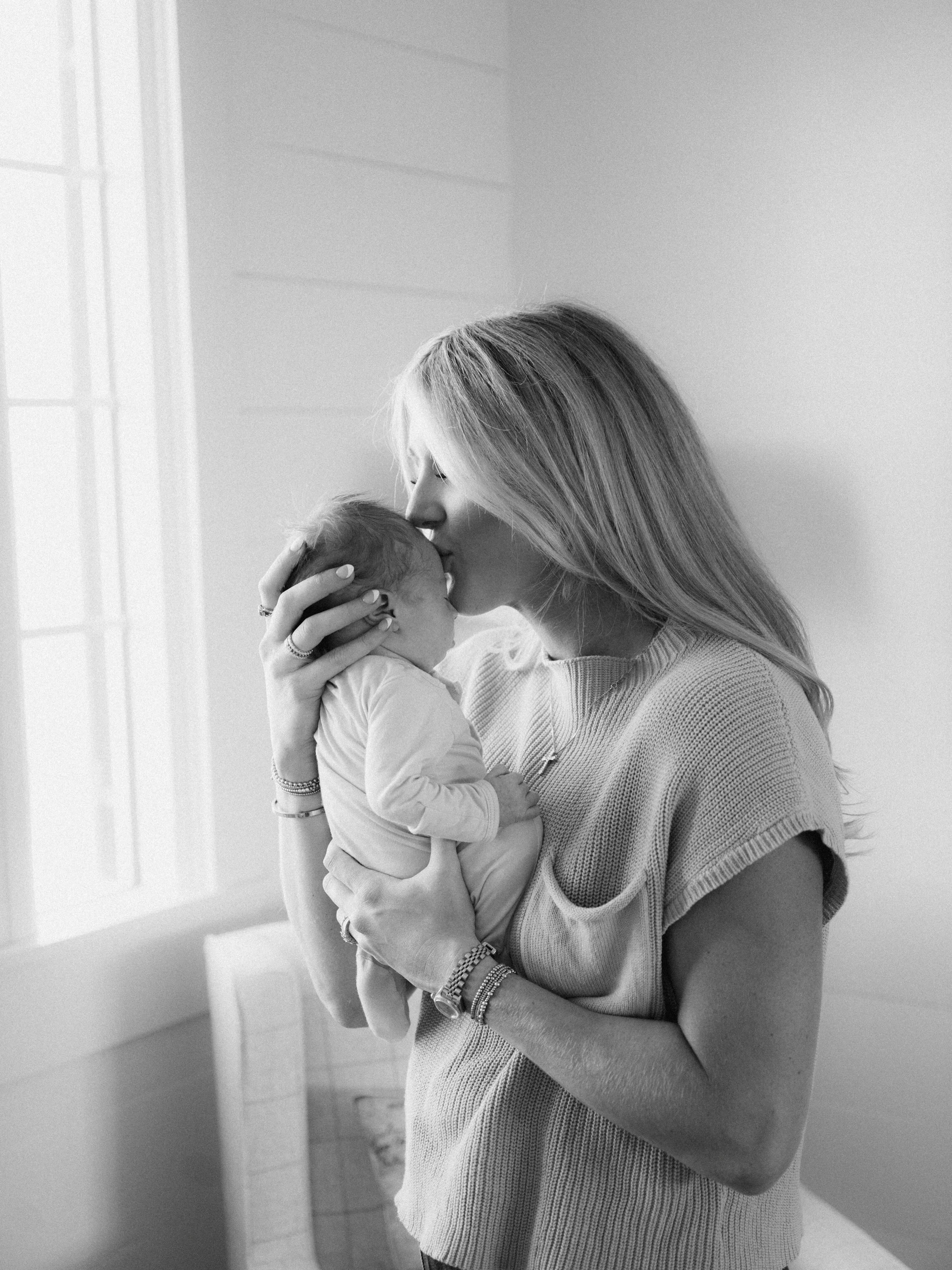 A black and white photo of a woman with long hair holding and kissing a newborn baby's head.
