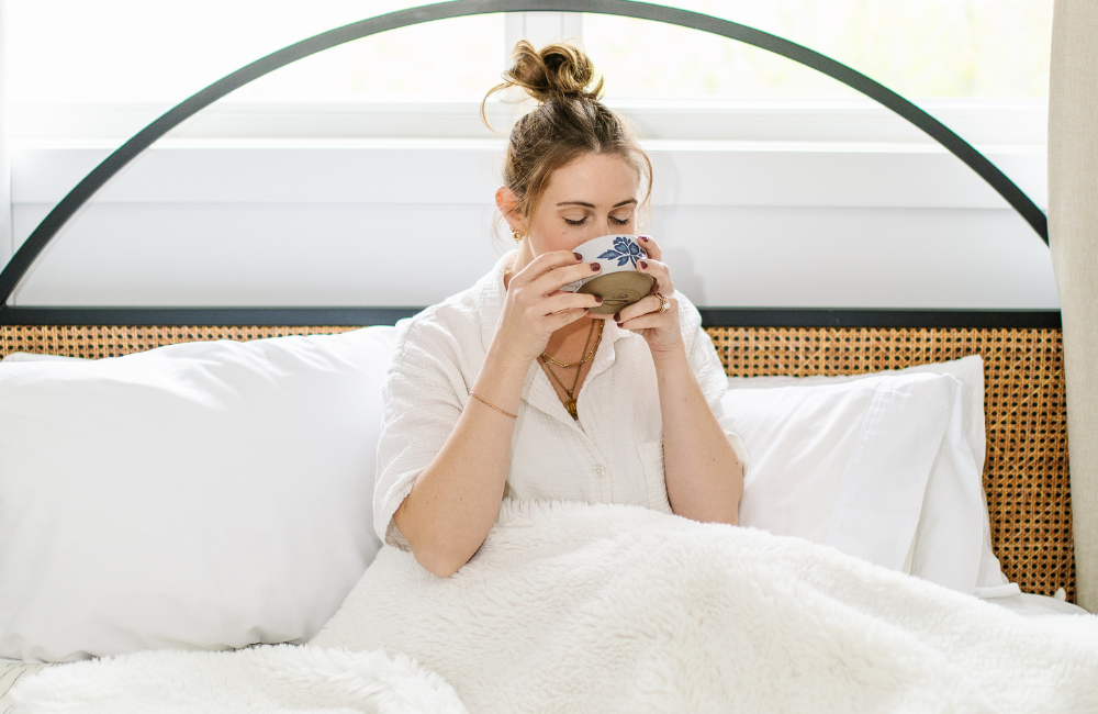 A woman with her hair in a top knot sits up in bed and drinks from a bowl.