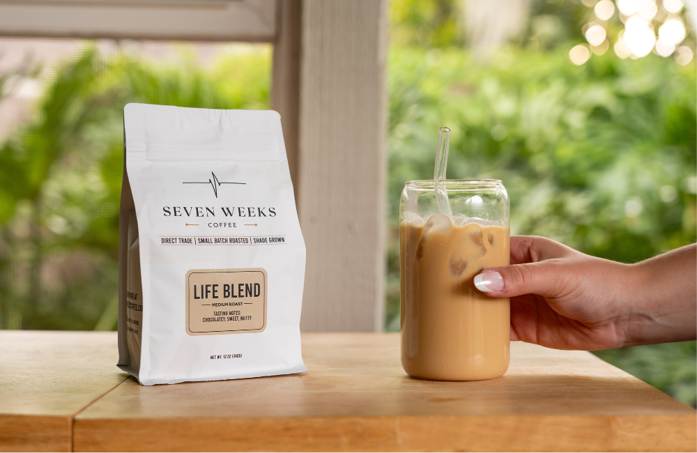 A hand holding a glass of iced coffee next to a bag of Seven Weeks Coffee on a table.