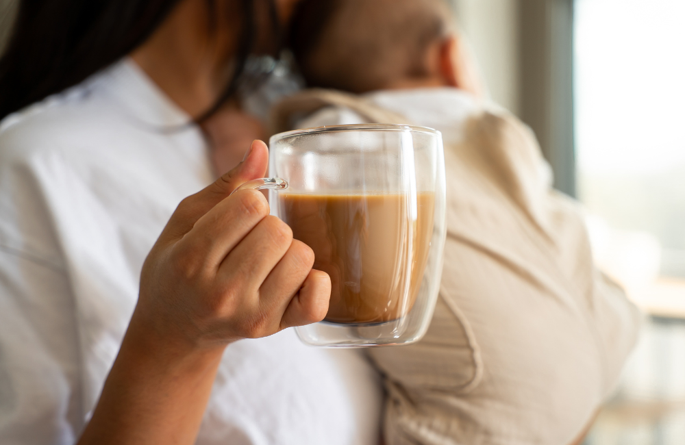 A person holds a clear glass mug with a brown beverage while holding a baby.