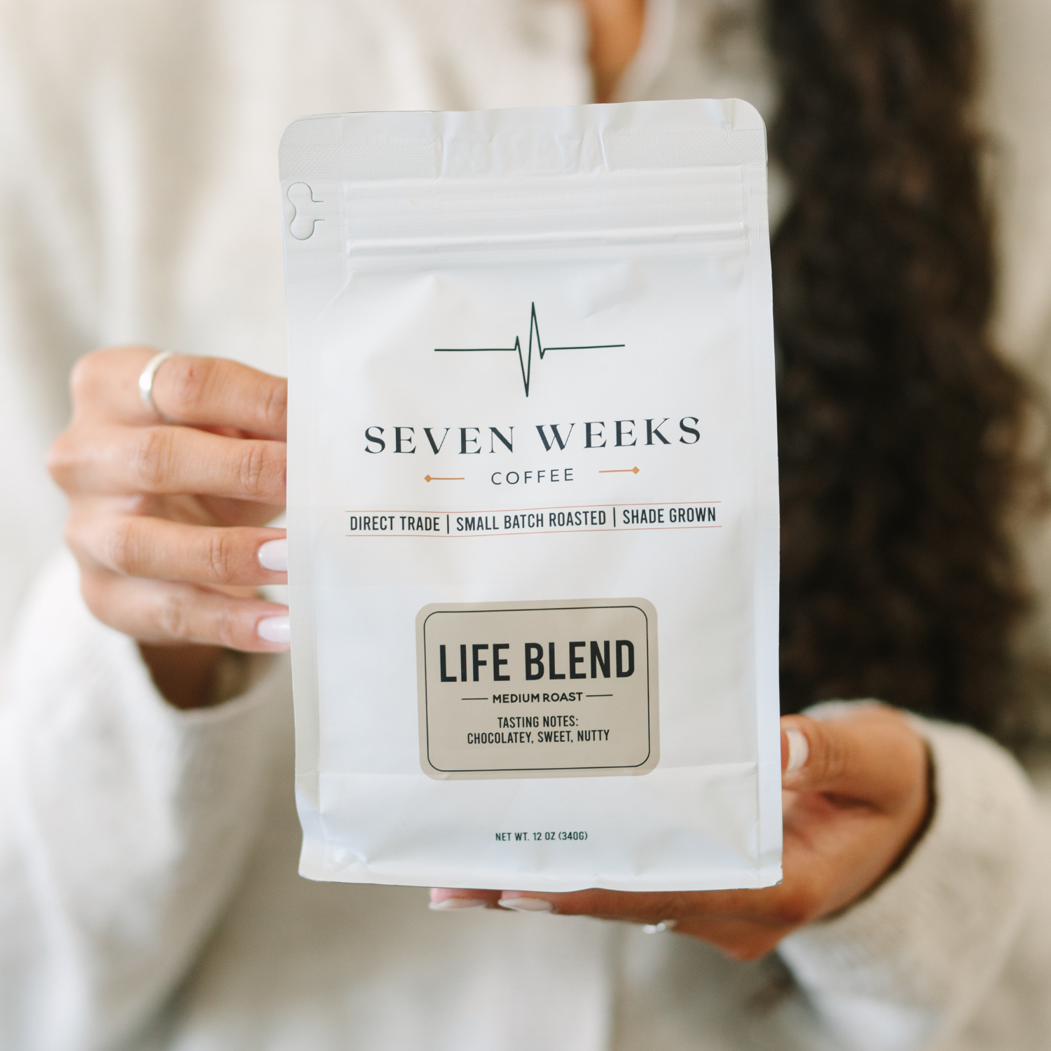 A person wearing a white sweater holds up a white bag of Seven Weeks Coffee Life Blend.