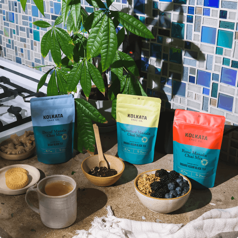 Three pouches of Kolkata Chai Co. mix on a counter with a cup of tea and breakfast items.