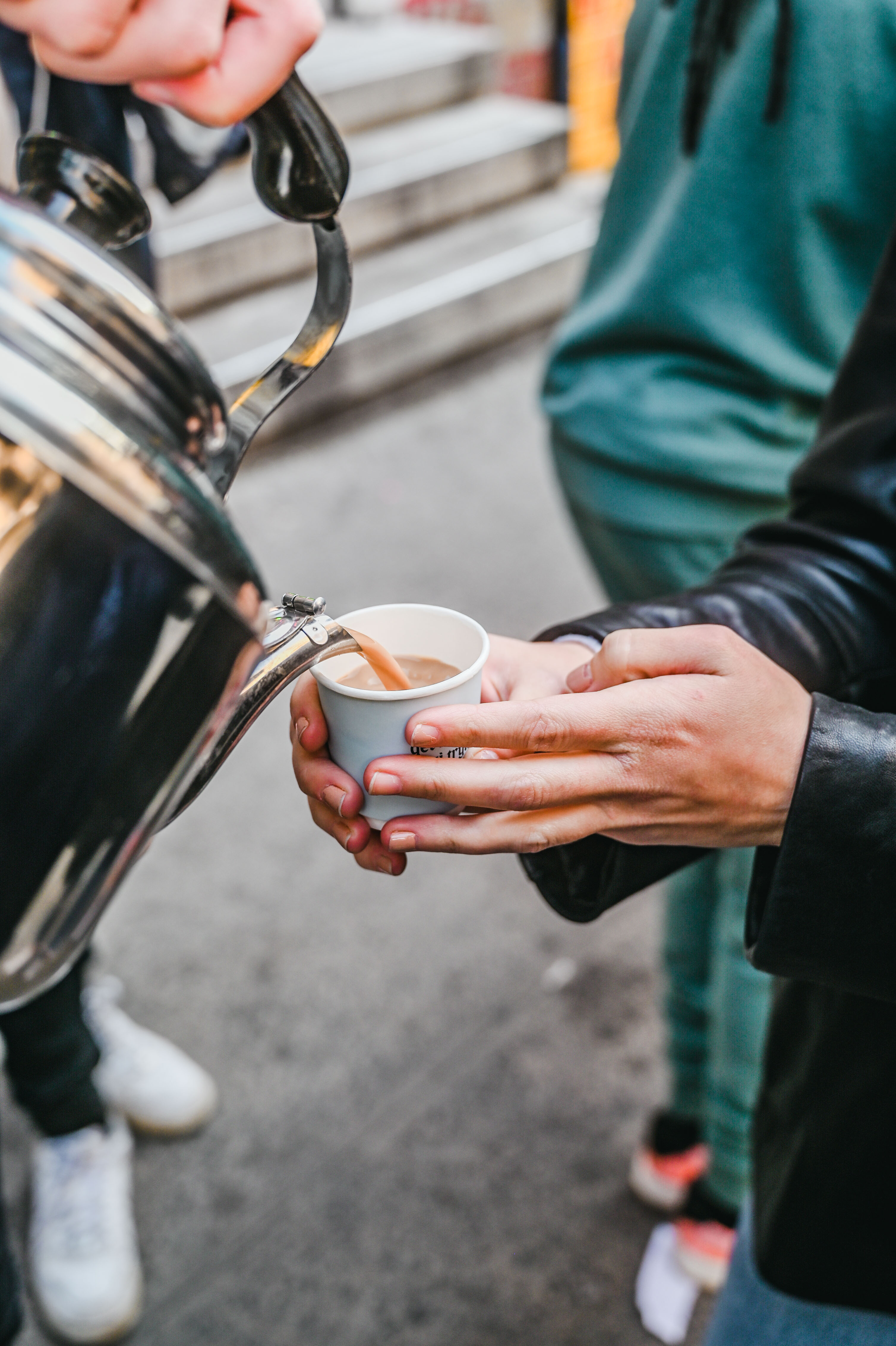 A person pours a beverage from a shiny metal urn into a paper cup held by another person.