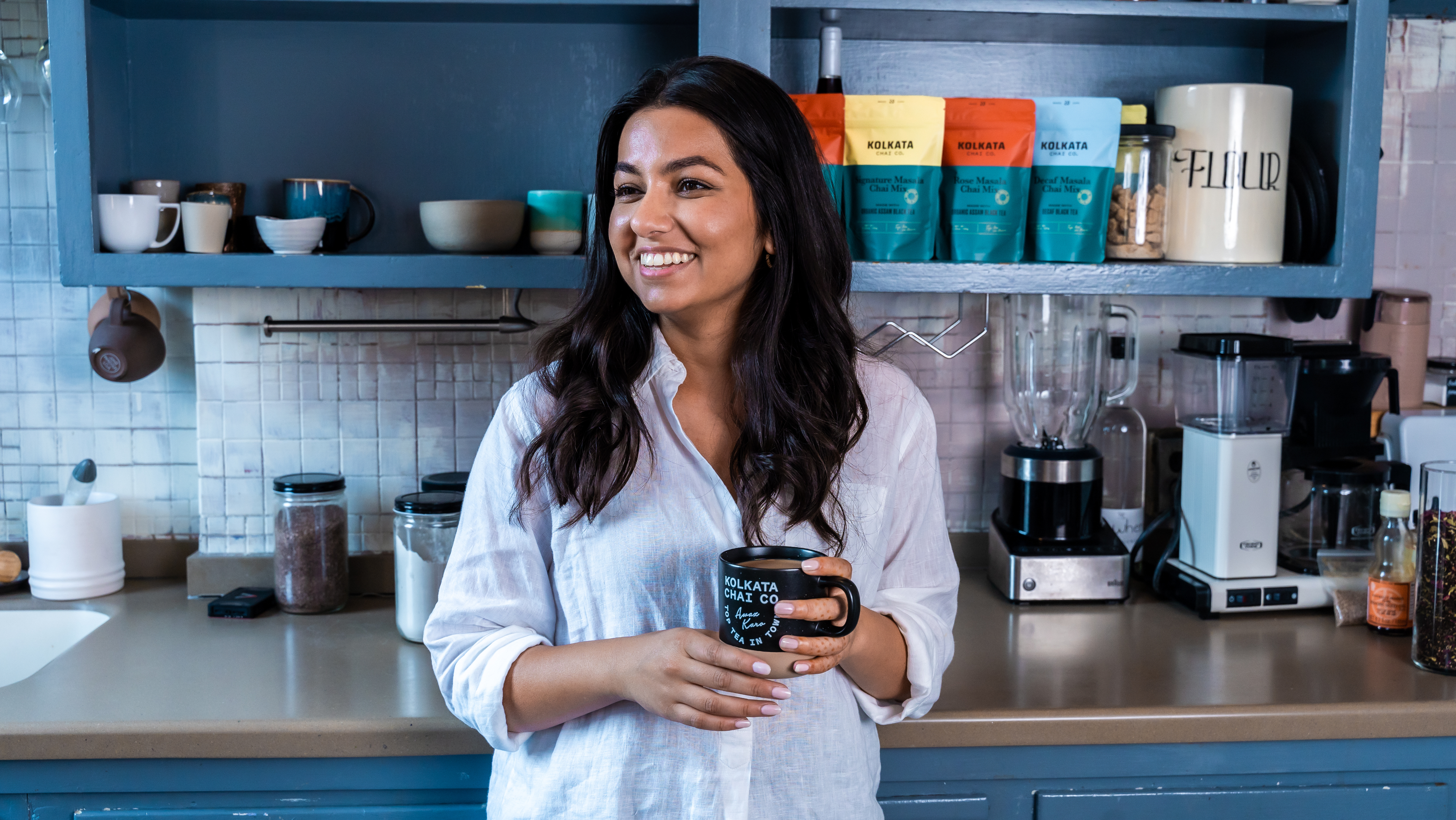 Smiling woman holding a mug in a kitchen with shelves and appliances.
