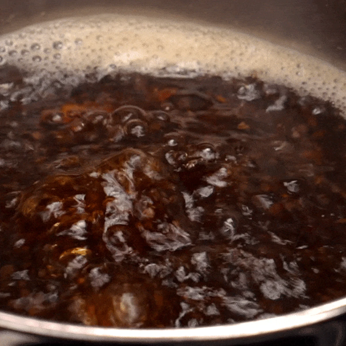 A close-up of a dark liquid boiling vigorously in a pot, creating bubbles and foam.
