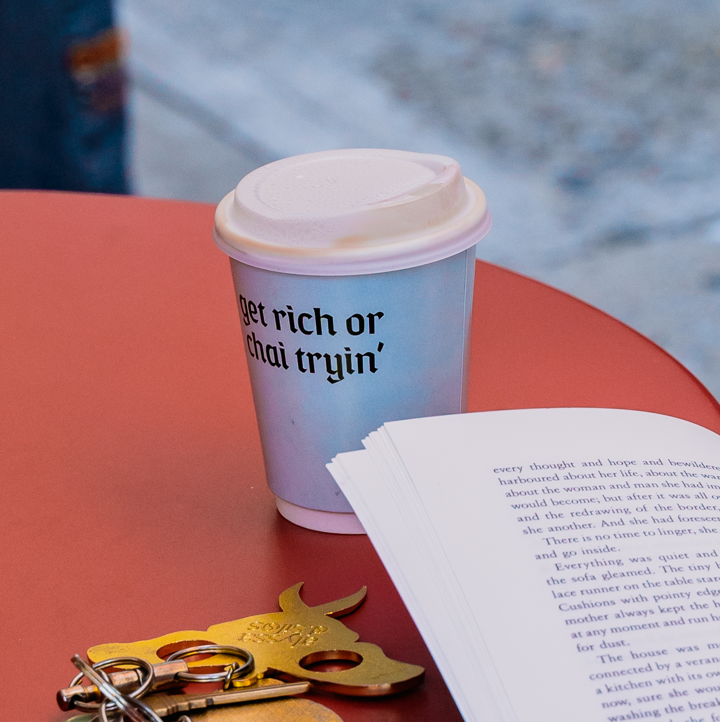 A takeaway coffee cup that reads 'get rich or chai tryin'' sits on a red table with a book and keys.