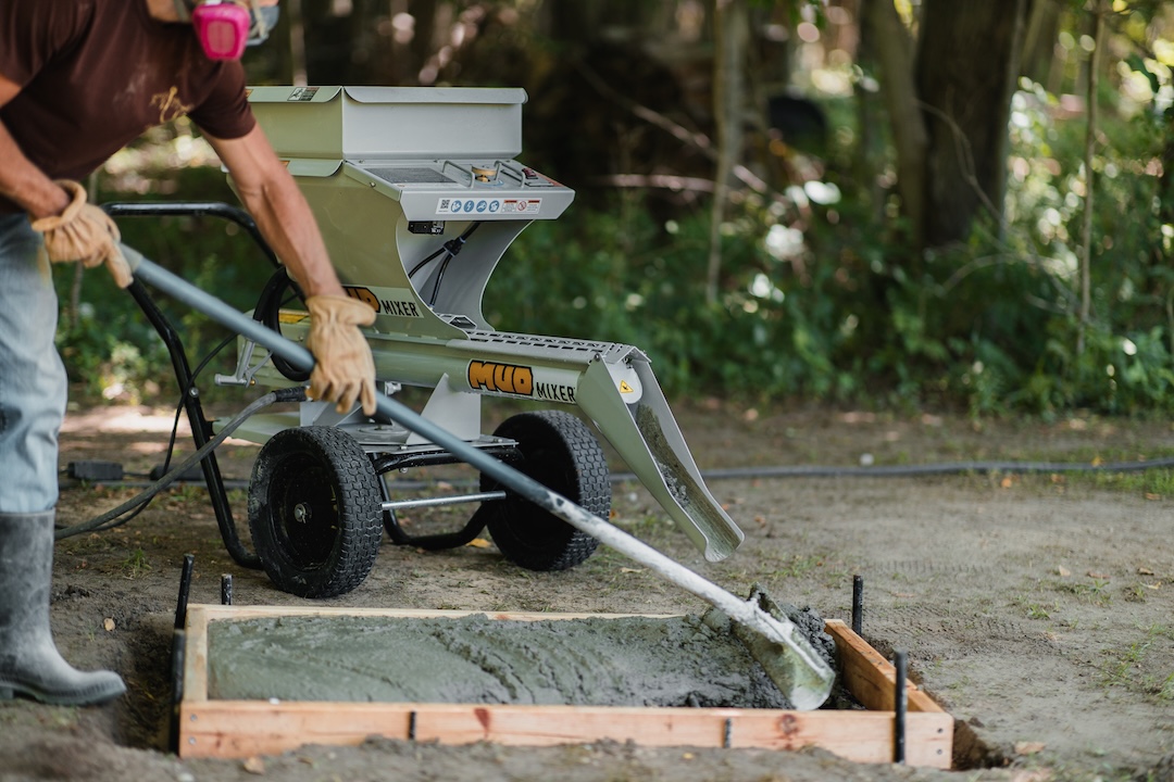 A worker pours concrete from a Mud Mixer machine into a wooden form on the ground.