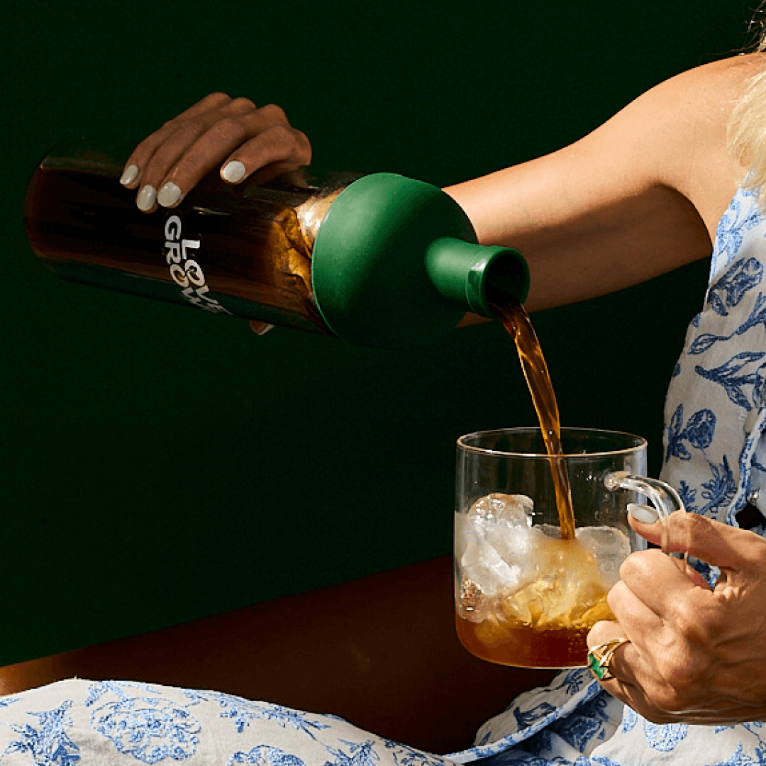 A person pours a dark beverage from a bottle with a green top into a clear glass mug of ice.
