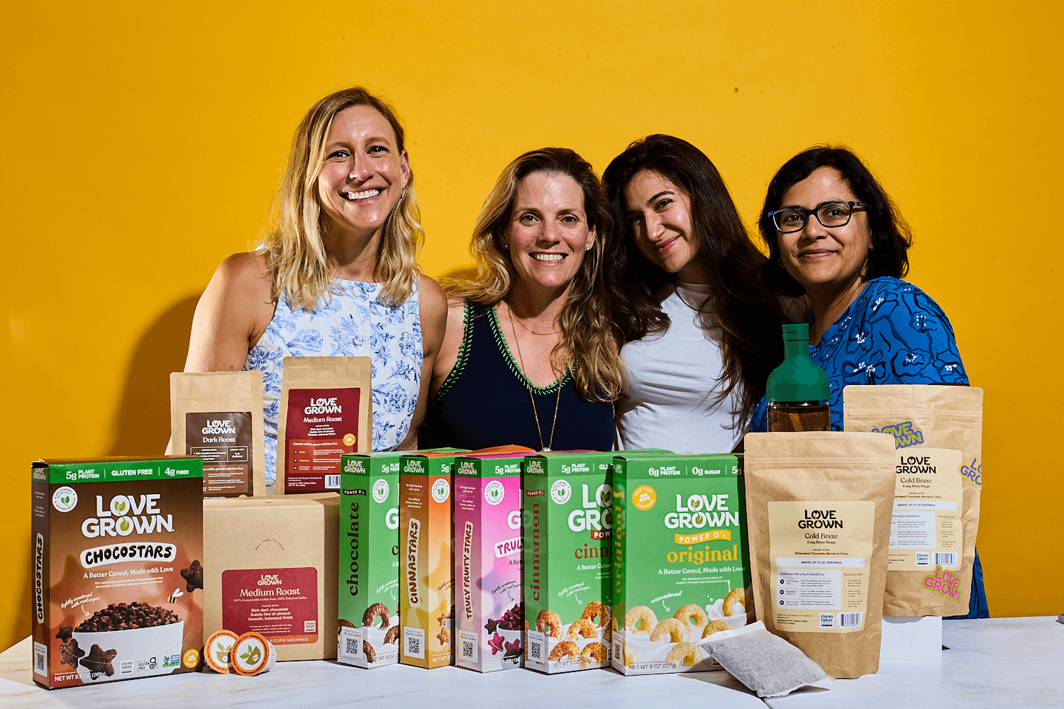 Four women smiling behind a table displaying various Love Grown cereal and coffee products against a yellow background.