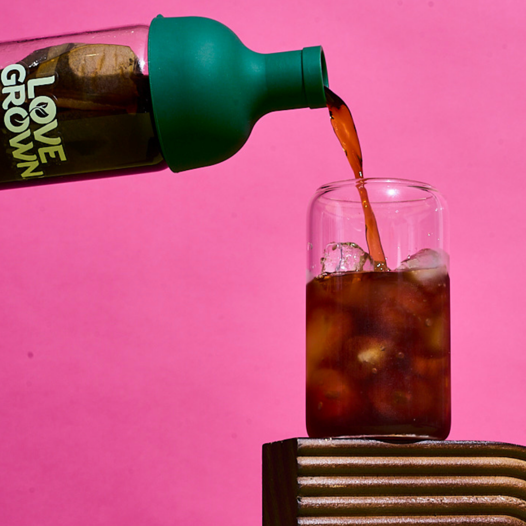 Iced tea is poured from a green-topped bottle into a glass of ice against a pink background.