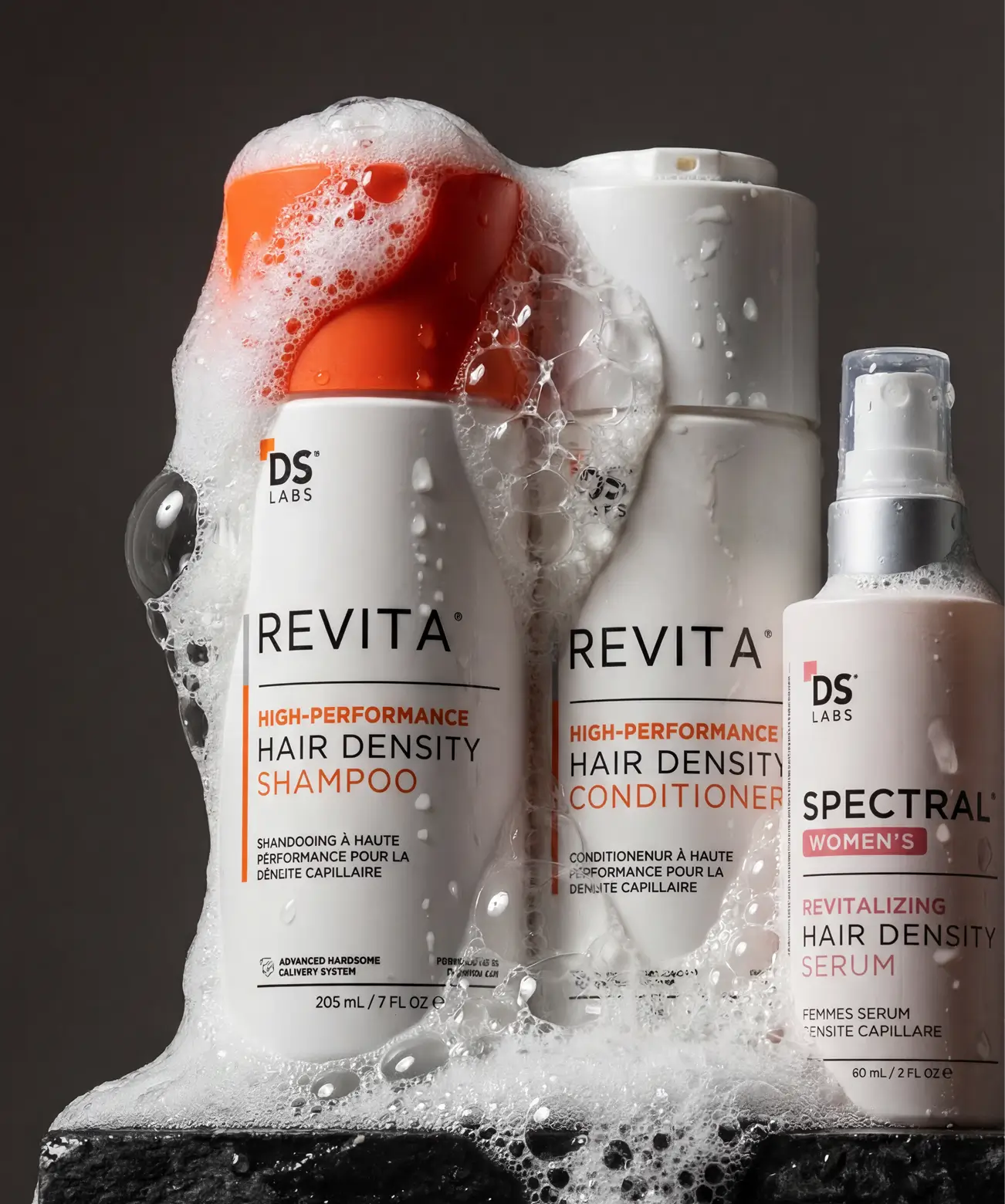 Three DS Labs hair care bottles—shampoo, conditioner, and serum—are covered in white suds against a dark background.