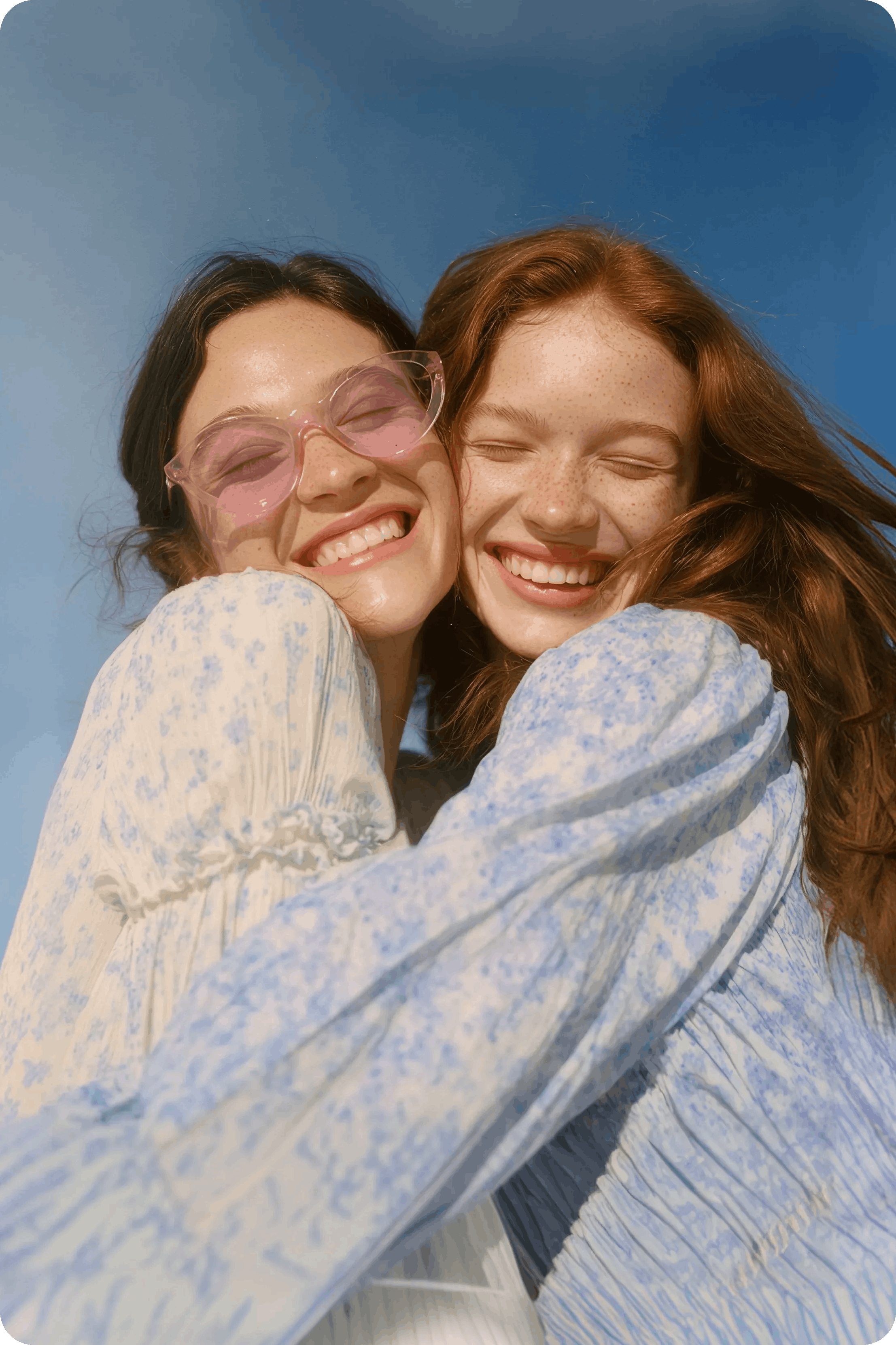 Two smiling young women in floral dresses hug each other warmly under a clear blue sky.