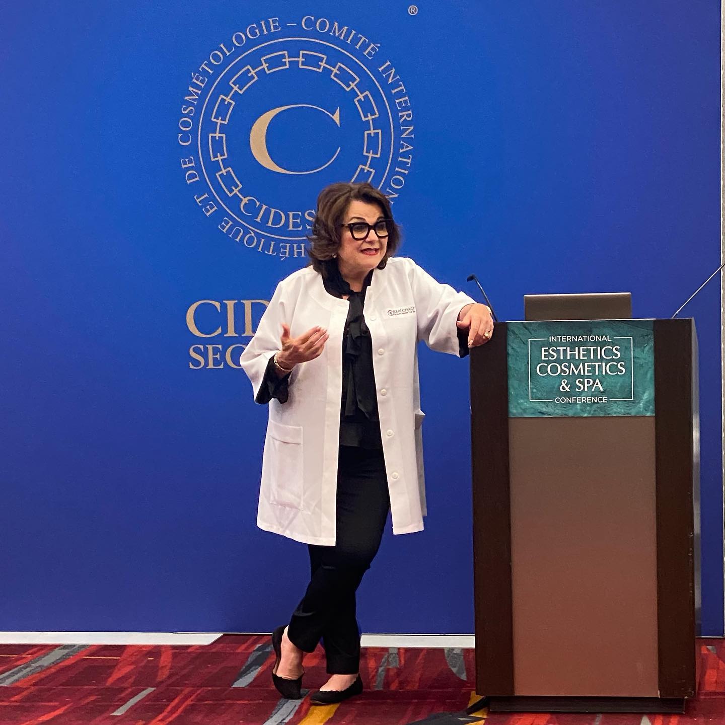 A woman in a white lab coat stands at a podium, speaking at a conference.