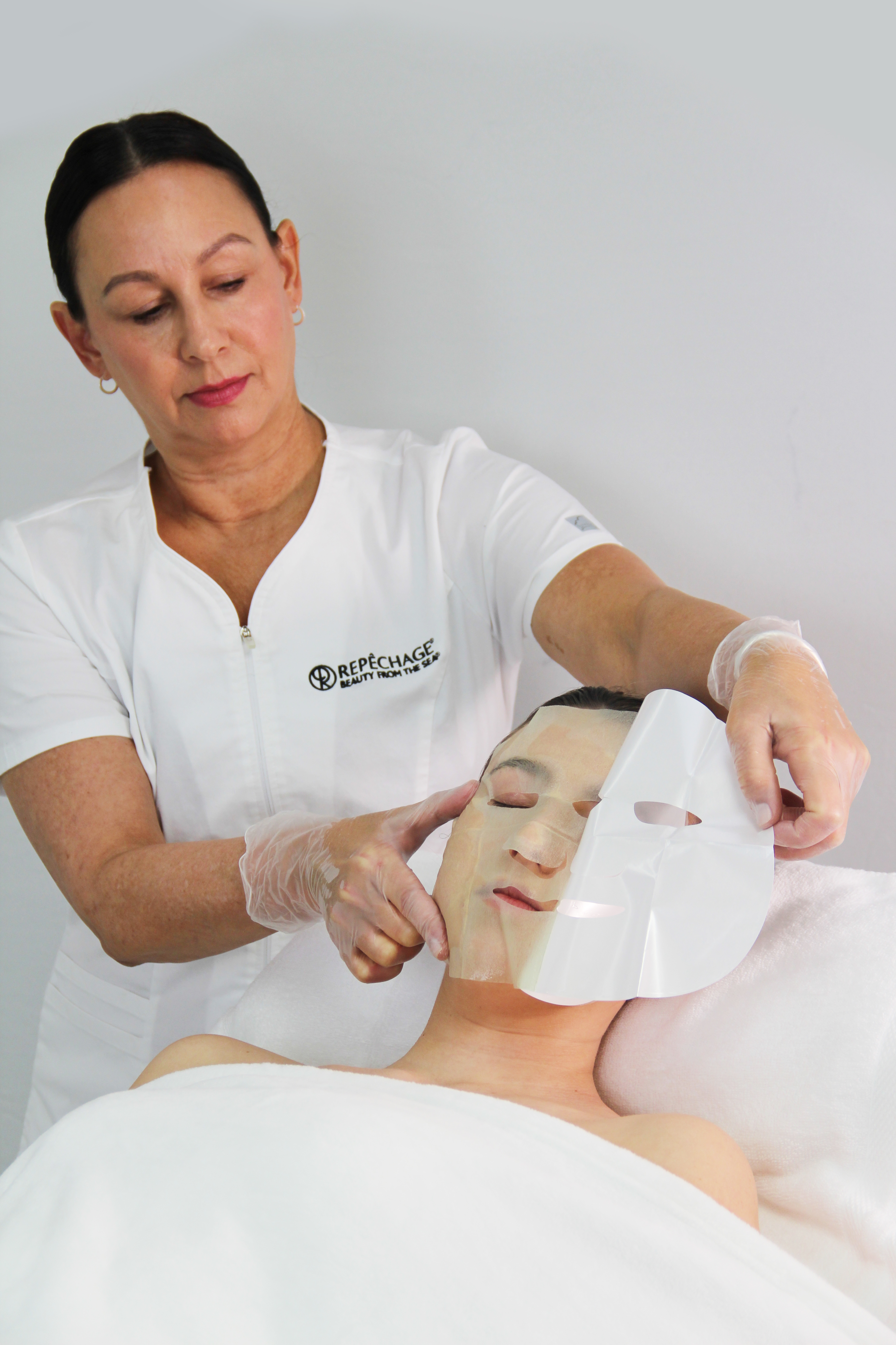 An esthetician in a white uniform applies a sheet mask to a client's face.