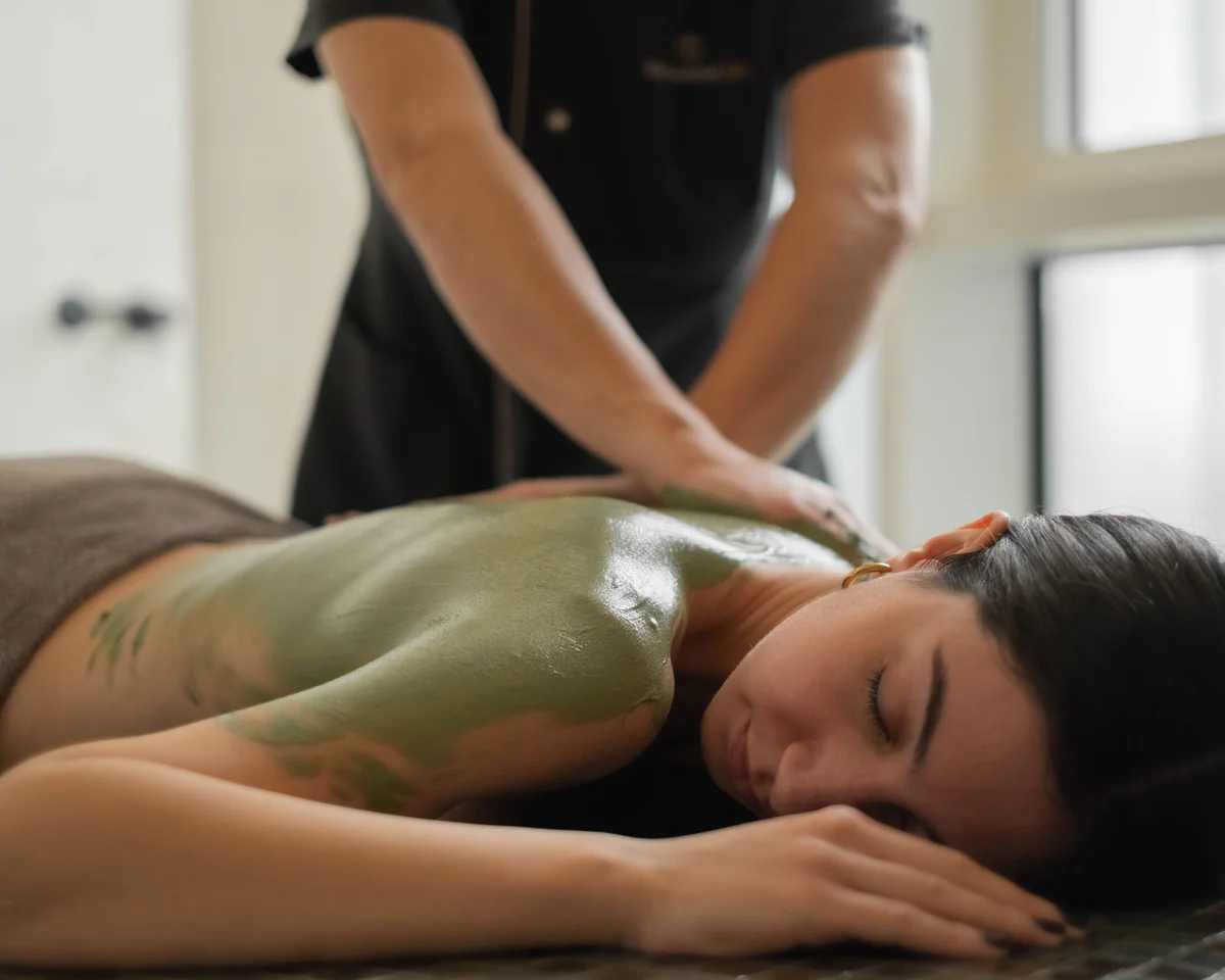 A woman lies on a massage table while a therapist applies a green body treatment to her back.