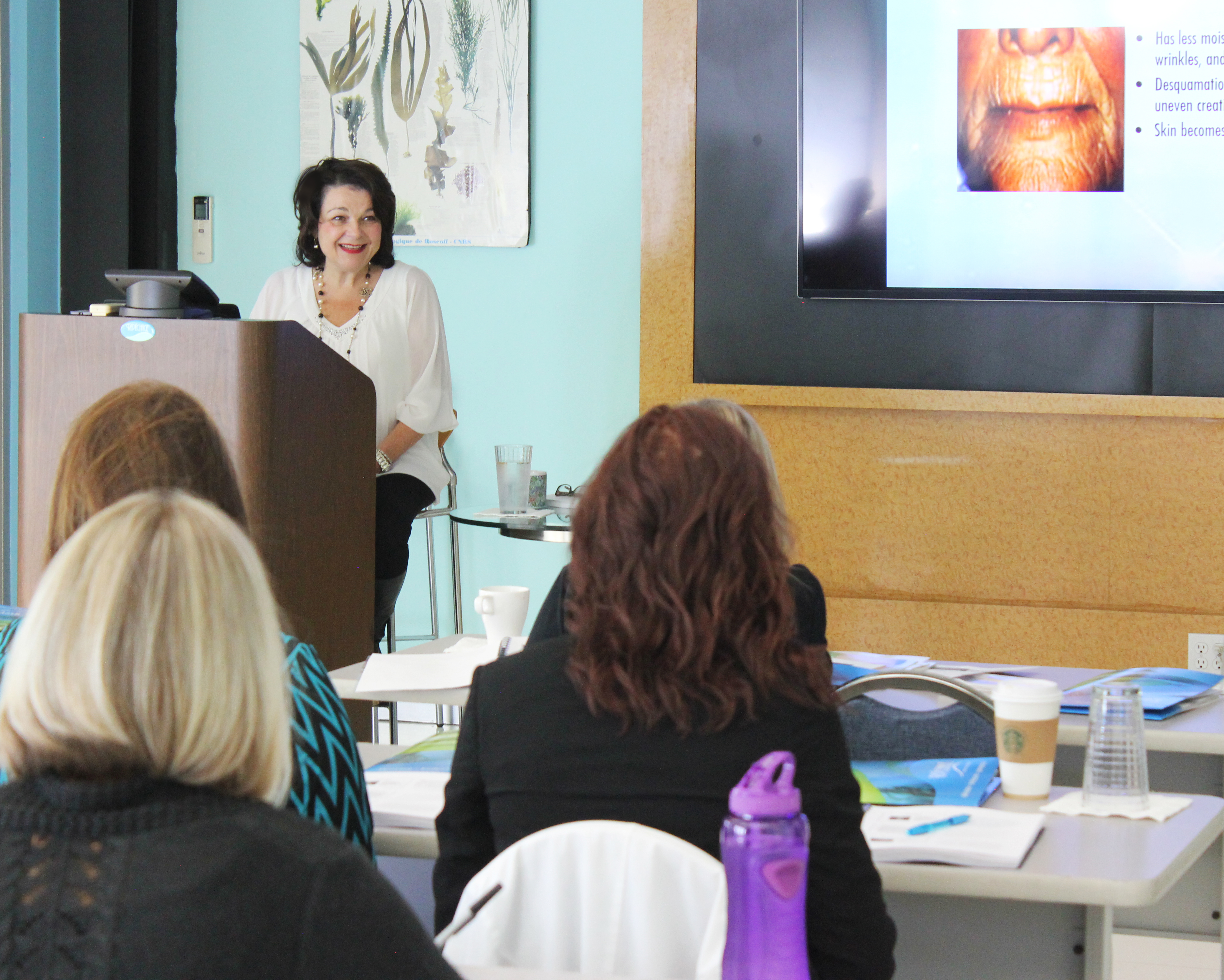 A woman stands smiling behind a podium, giving a presentation to a seated audience in a classroom.