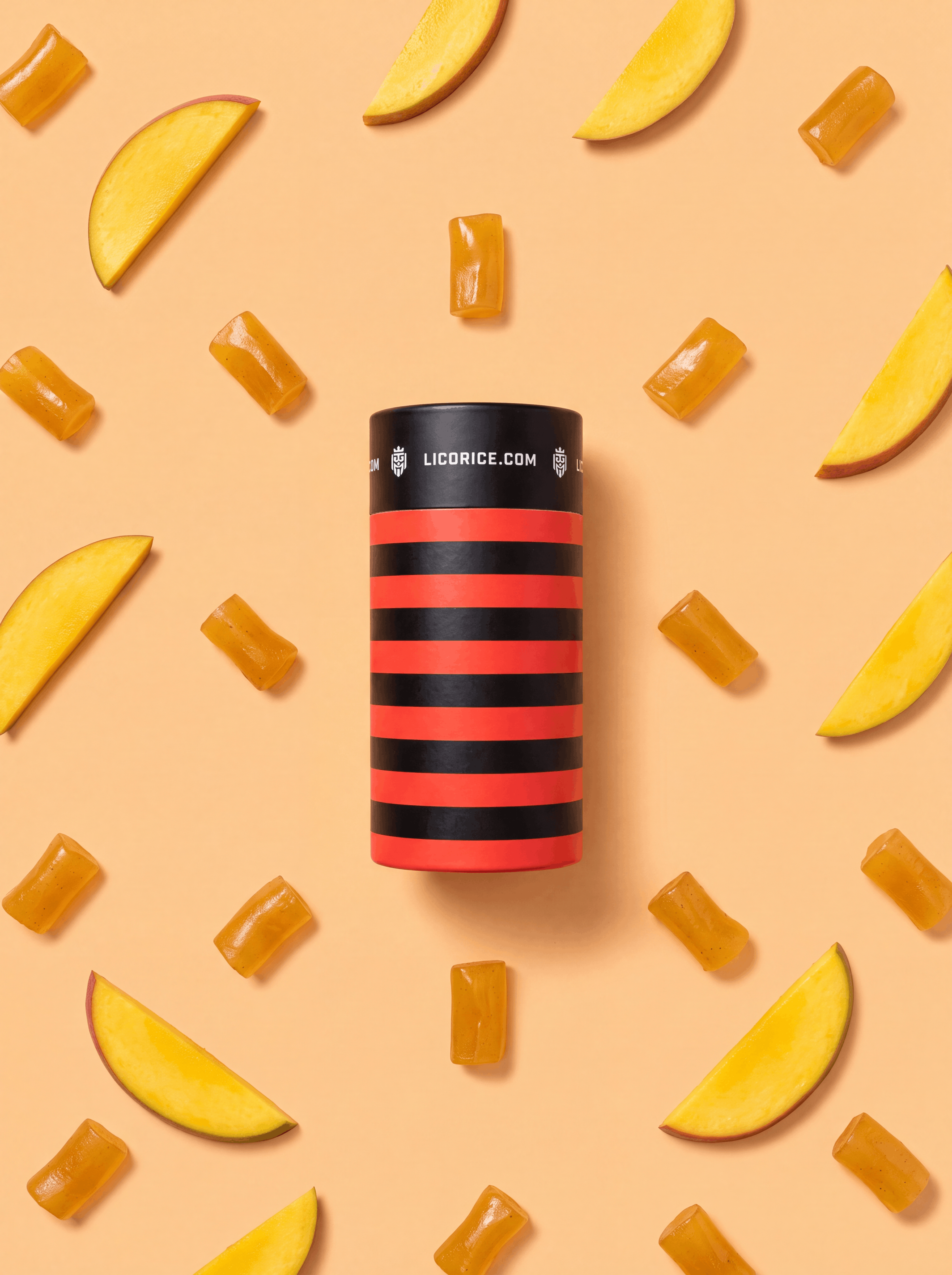 A black and red striped licorice container is surrounded by mango slices and candies on a peach background.