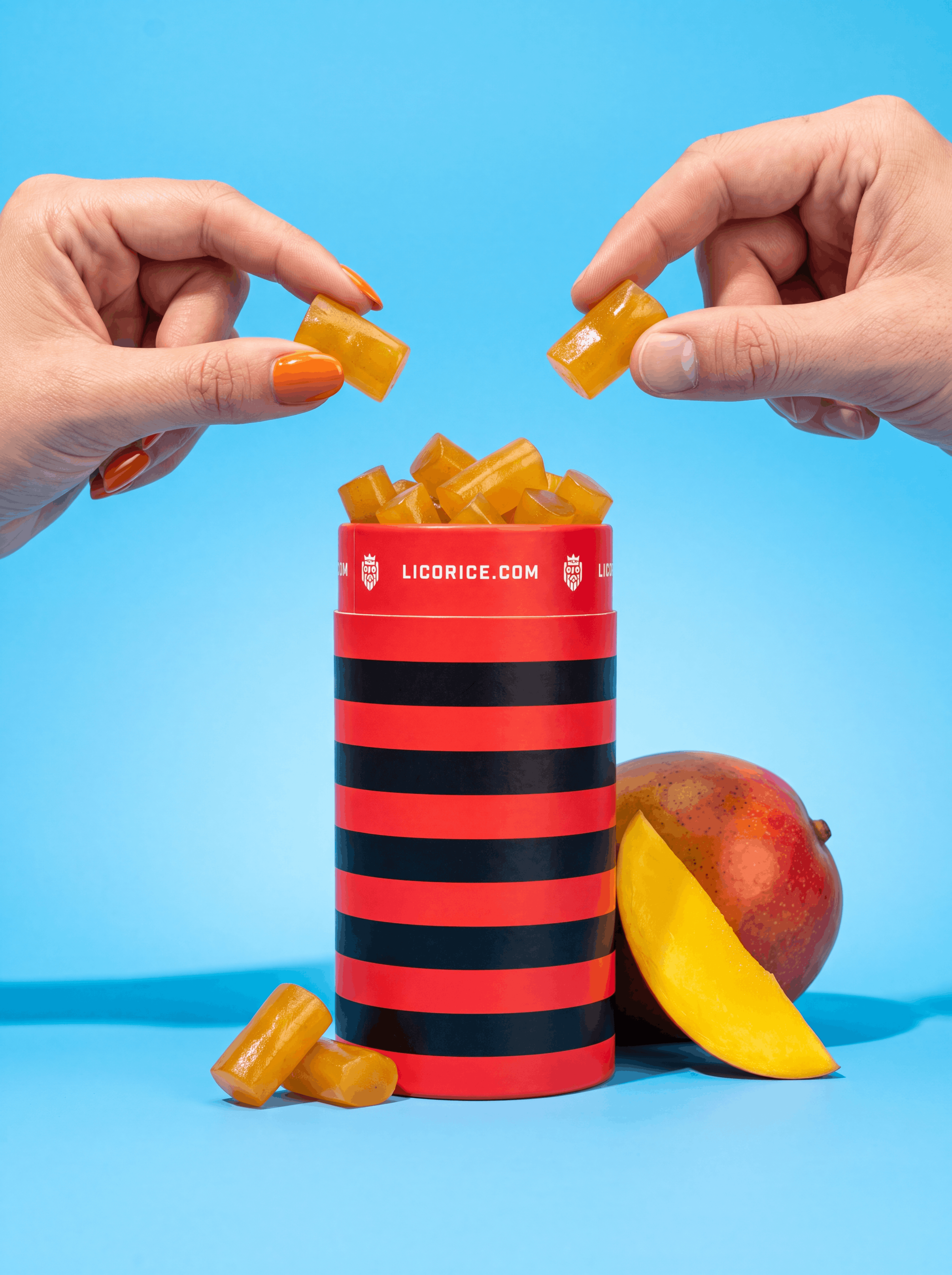 Two hands hold yellow licorice pieces over a striped container next to a sliced mango against a blue background.