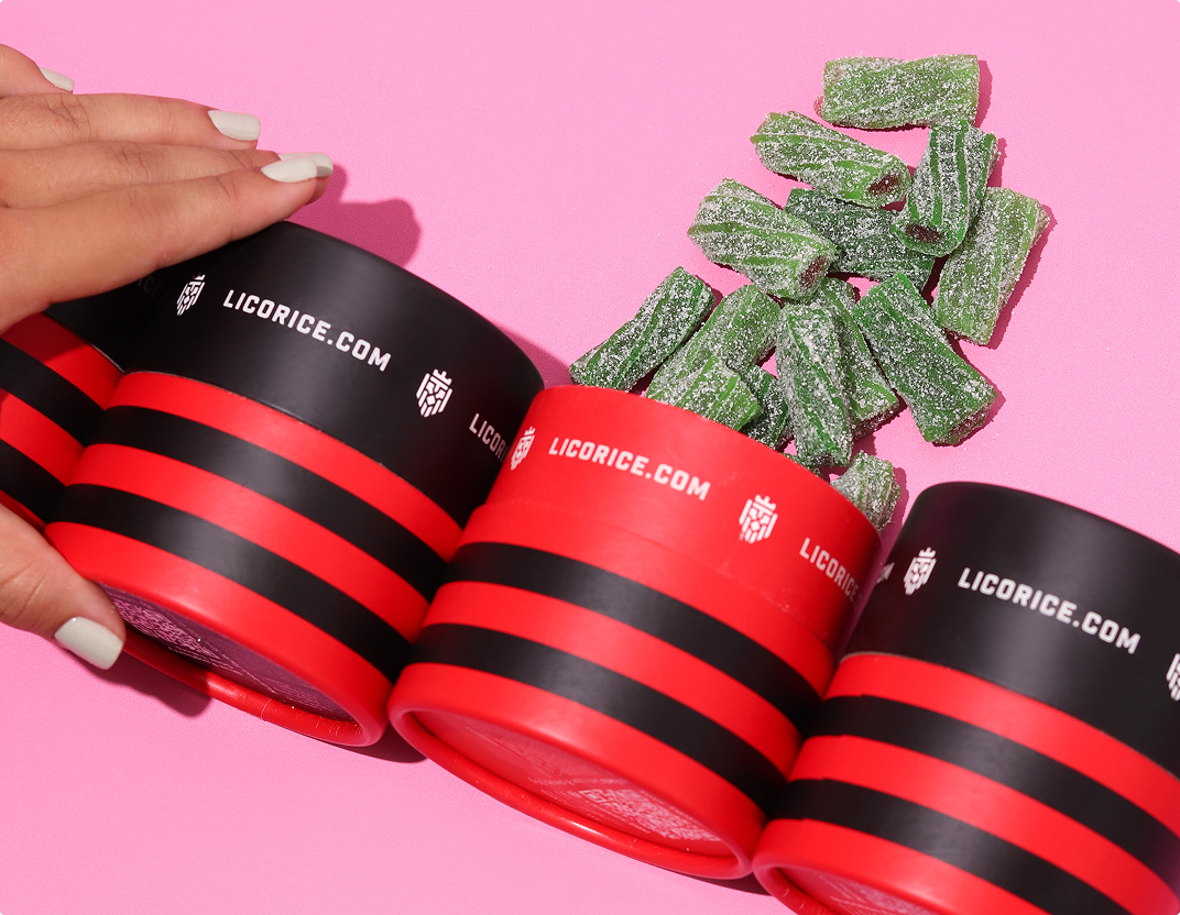 A hand reaches for red and black striped containers of green, sugar-coated licorice on a pink background.