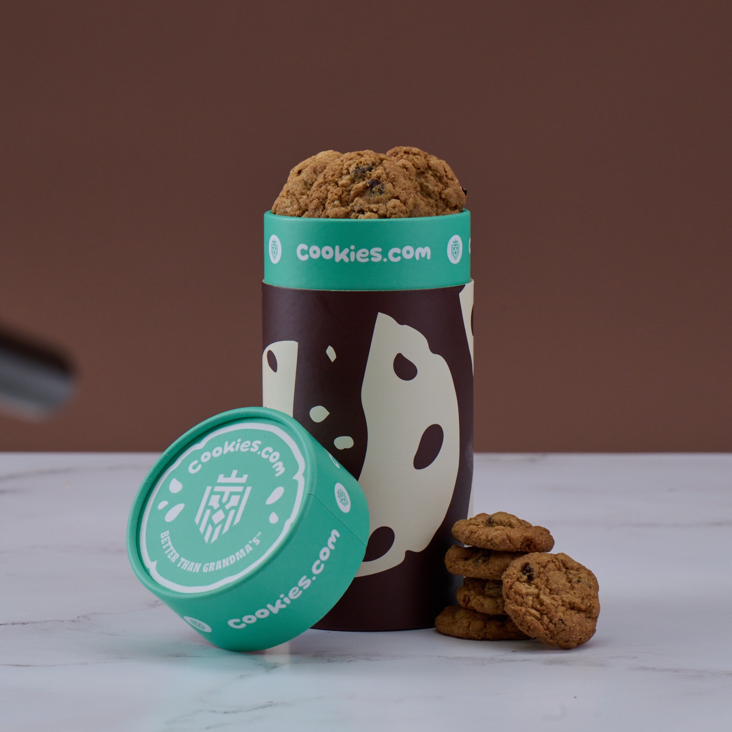 A cylindrical cookie container with cookies inside and beside it, labeled cookies.com.