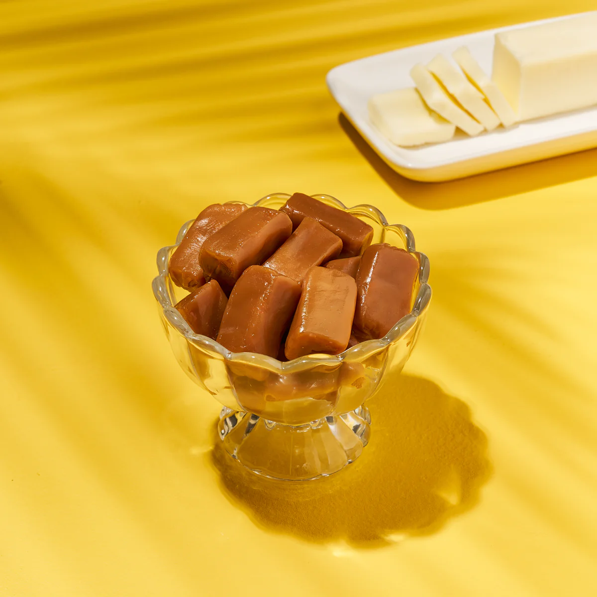 Glass bowl with caramel candies beside sliced butter on yellow background.
