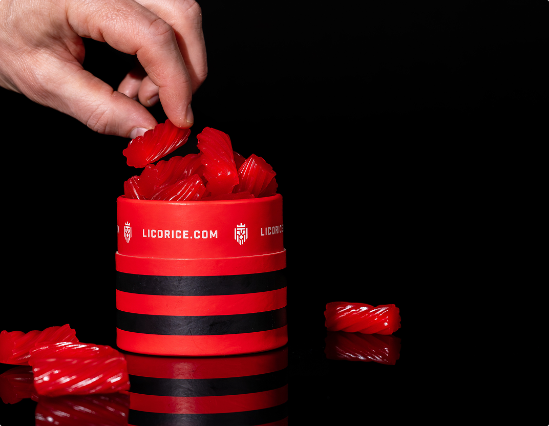 A hand takes a piece of red licorice from a red and black striped container against a black background.