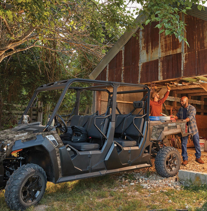Two people loading gear into the bed of a Tracker side-by-side parked outside a rustic barn