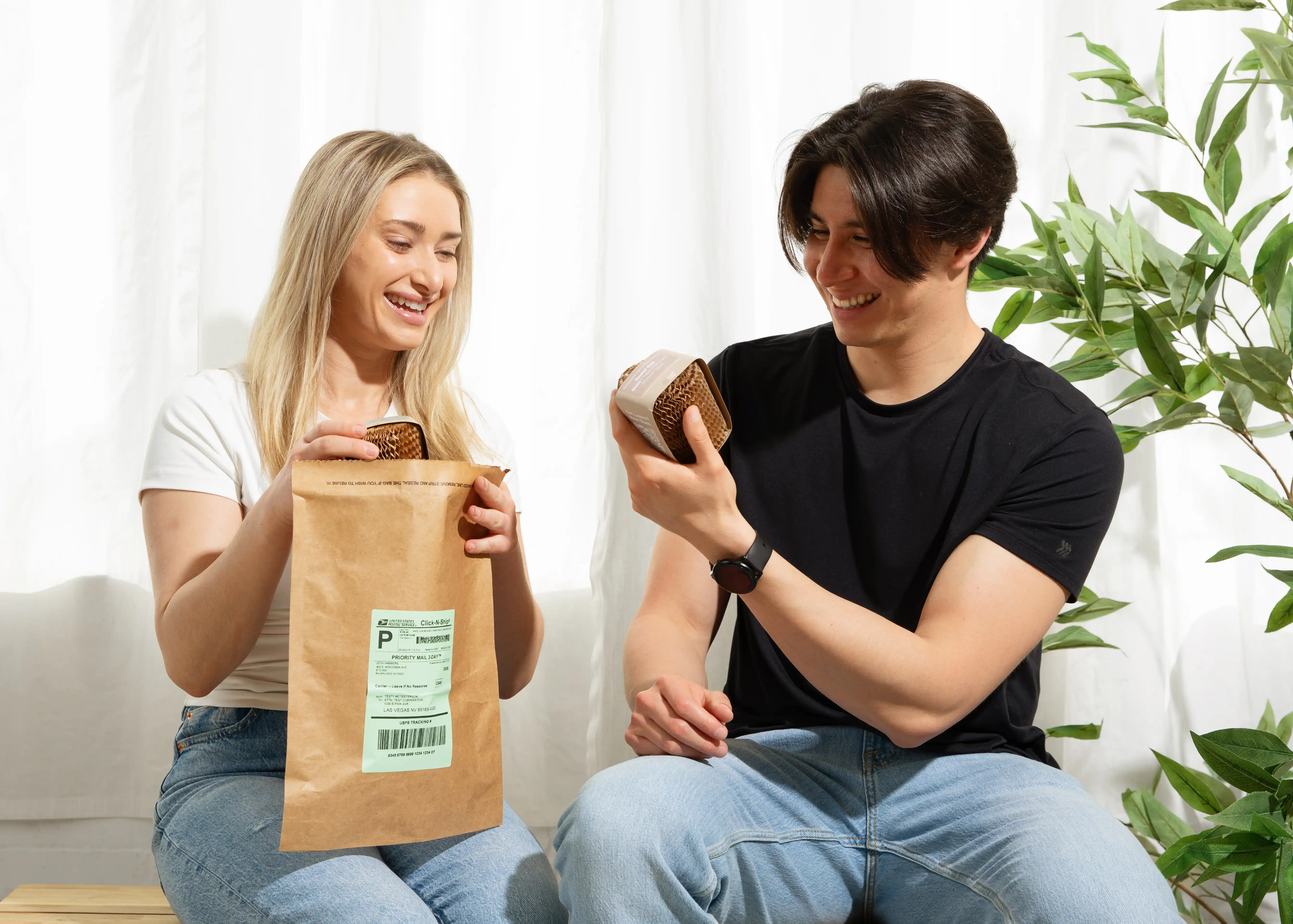 A smiling man and woman sit next to each other, unboxing a package with honeycomb paper wrapping.