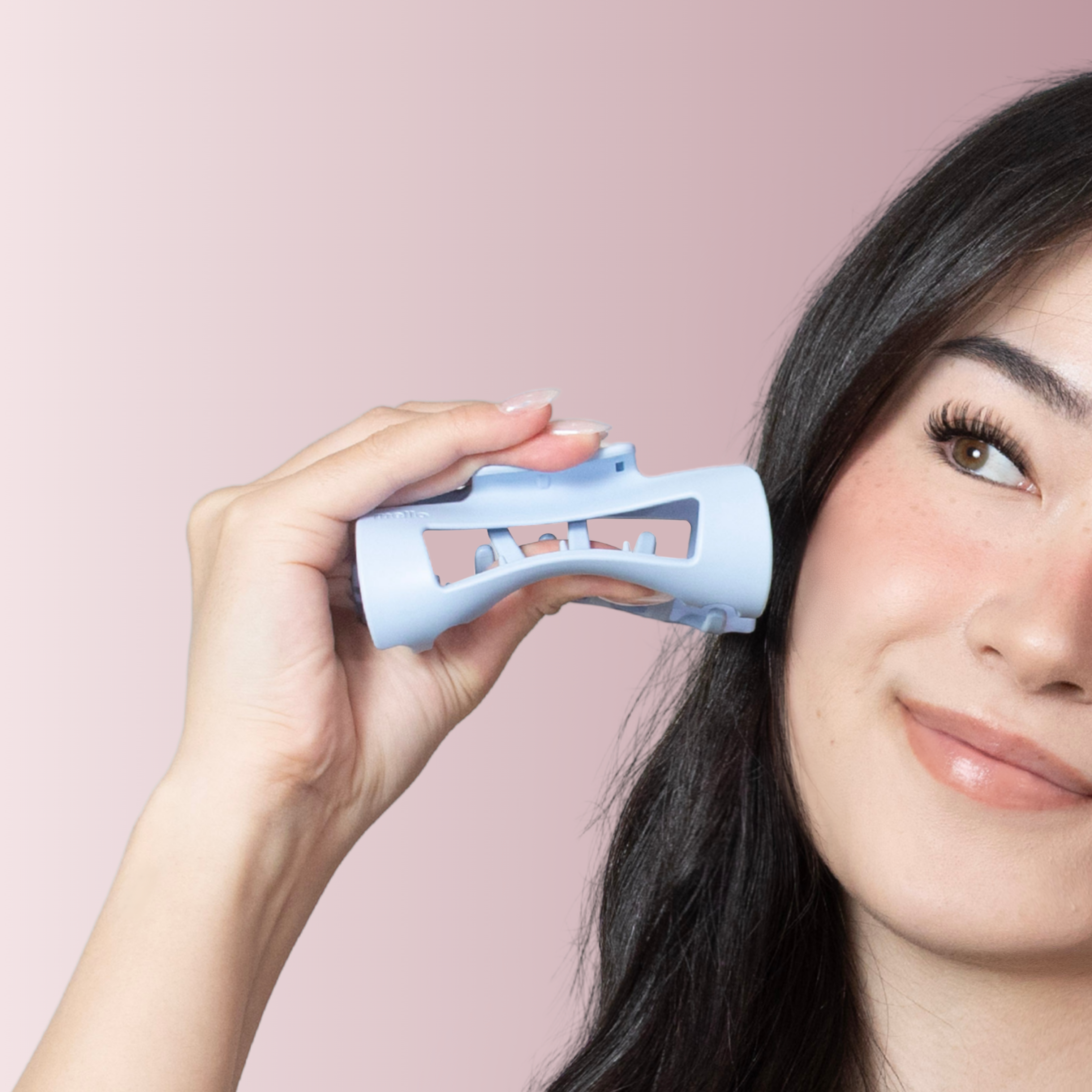 A woman with dark hair holds a light blue roller-style hair clip up to her head and smiles.