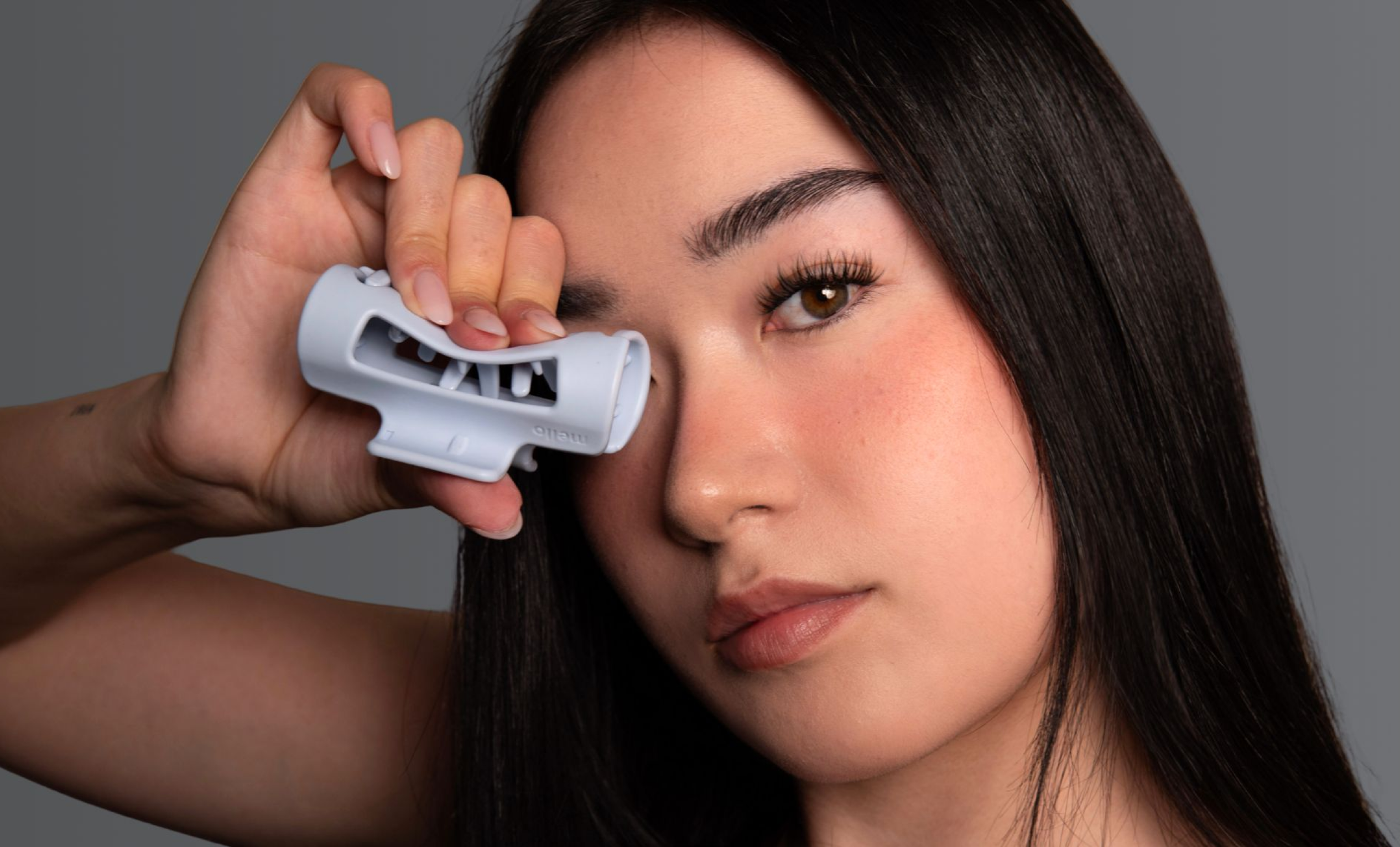 A woman with long dark hair holds a pale blue beauty tool over her right eye.