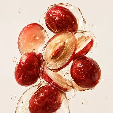 Whole and halved red plums are suspended in a splash of clear liquid against a cream background.