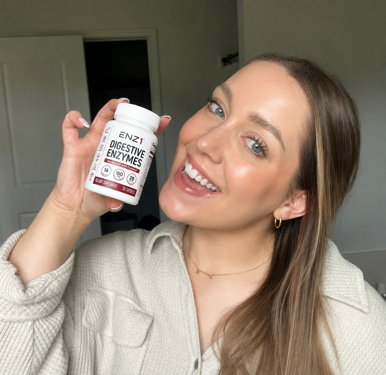 A smiling woman with blue eyes holds up a white bottle of ENZ1 Digestive Enzymes.