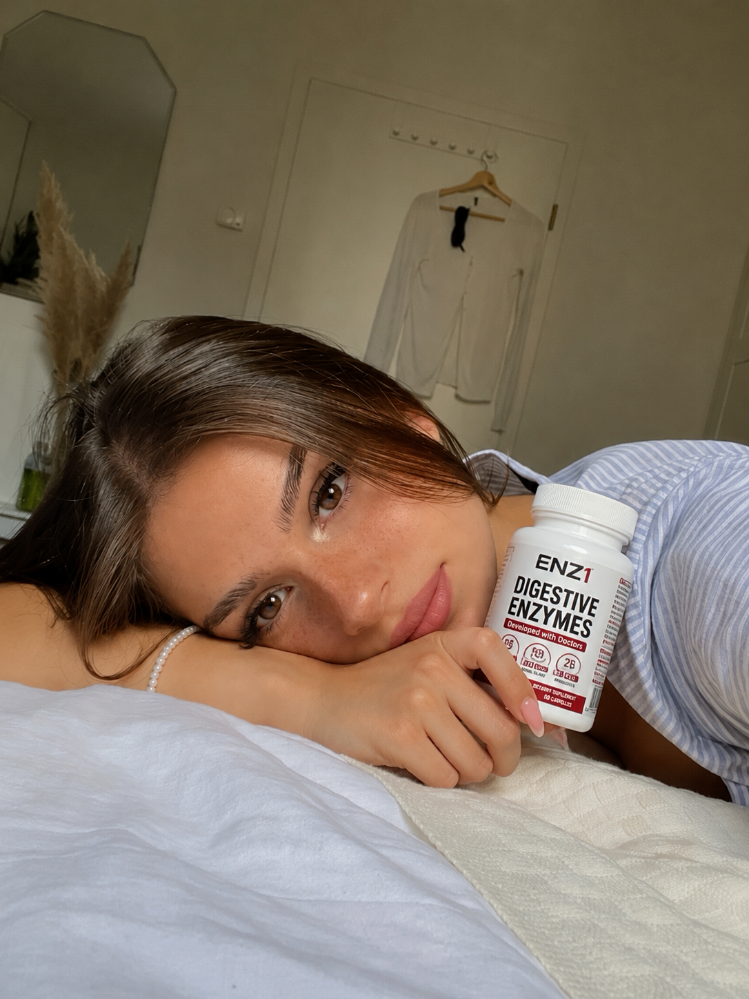 A woman lies on a bed, resting her head on her arm while holding a bottle of digestive enzymes.