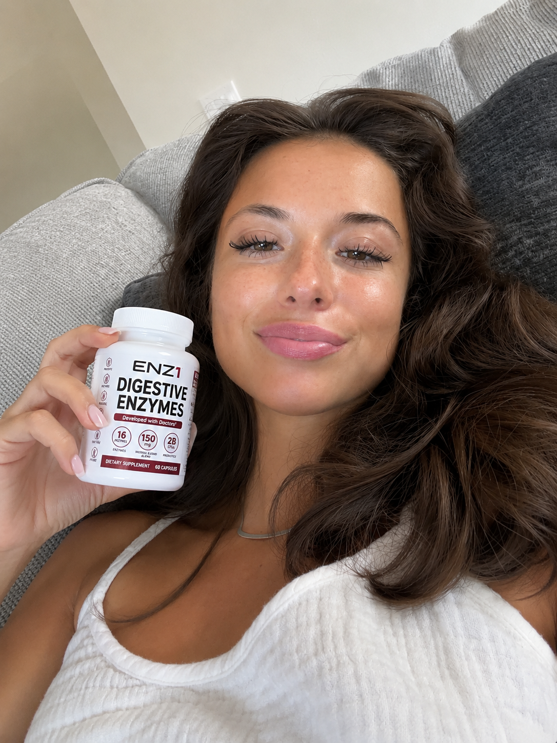 A woman lying on a couch holds up a white bottle of ENZ1 Digestive Enzymes.