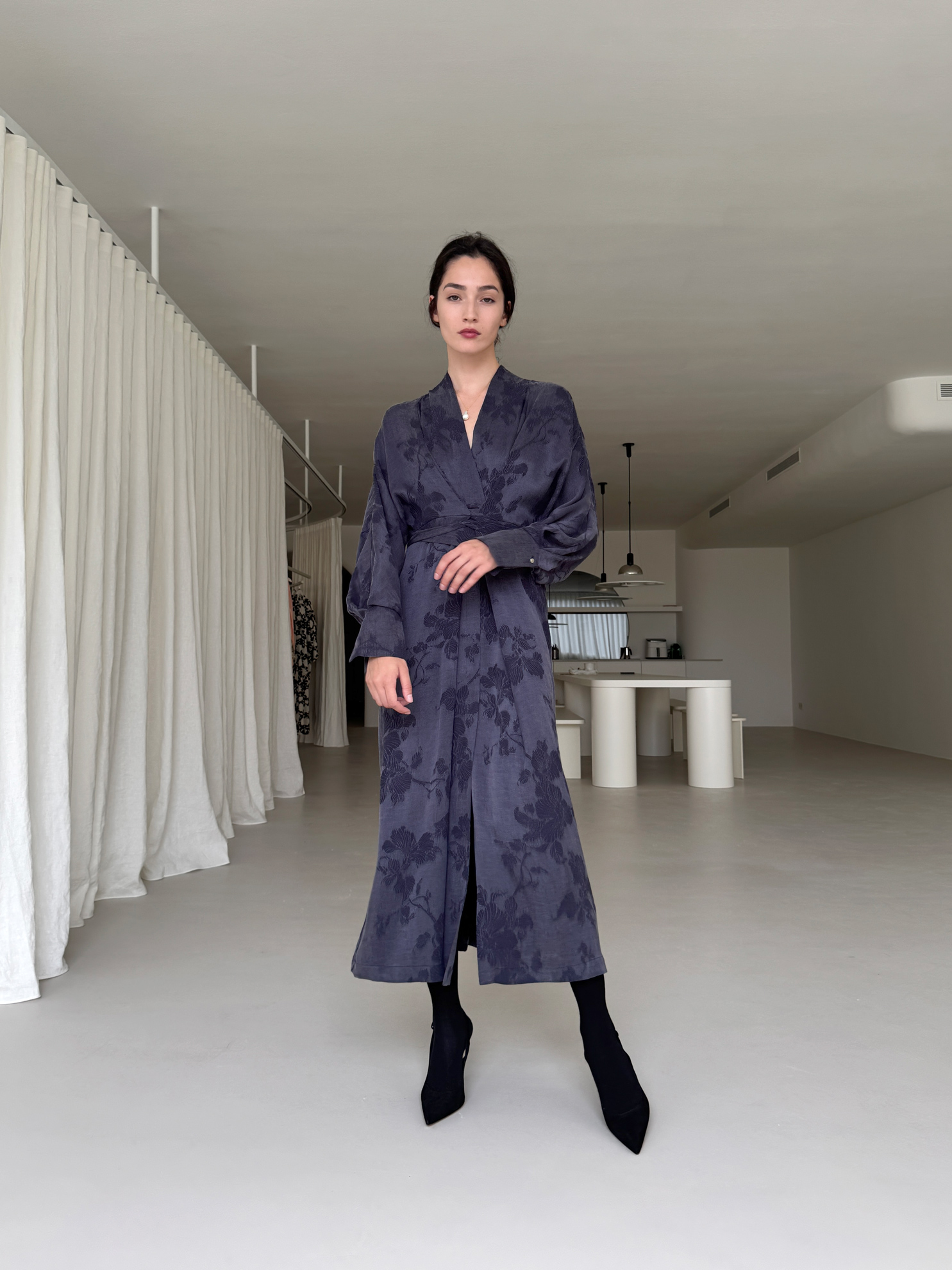 Person in a long, dark, patterned dress and black boots in a minimalist room.