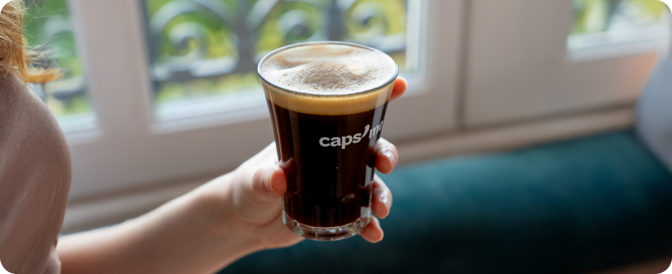 A person's hand holds a glass of dark, foamy coffee in front of a window.