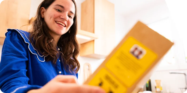 A smiling woman in a blue shirt looks down at a package in a kitchen.