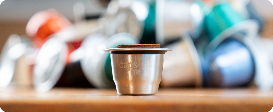 A reusable metal coffee capsule in focus, with a blurry pile of used, colorful disposable pods behind it.