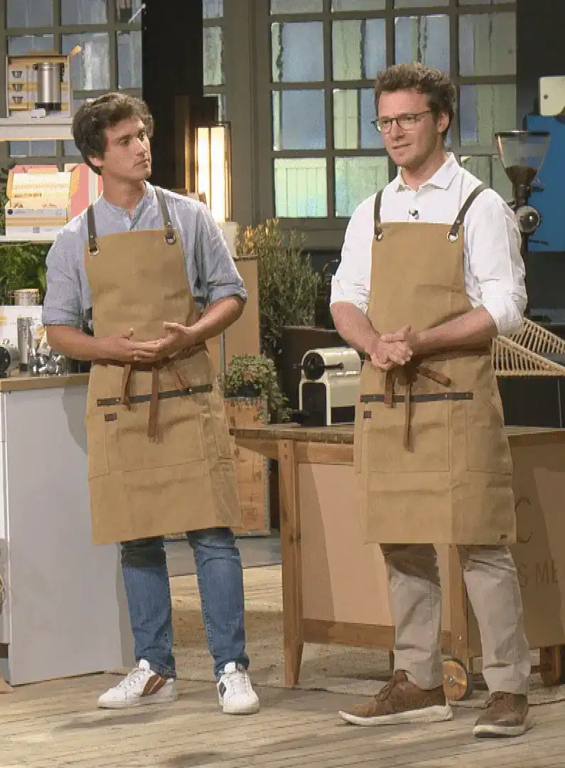 Two men wearing aprons stand in a workshop setting.