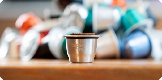 A reusable metal coffee capsule sits on a wooden table in front of a pile of used capsules.