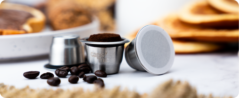 Reusable metal coffee pods, one filled with grounds, surrounded by coffee beans on a marble surface.