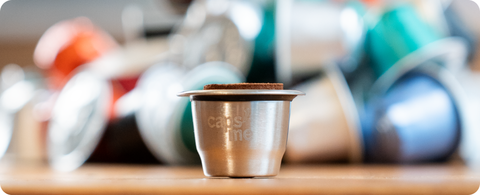 A reusable stainless steel coffee capsule filled with coffee grounds, with a pile of used capsules blurred in the background.