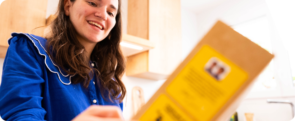 A smiling woman in a blue shirt looks down at a package in a kitchen.