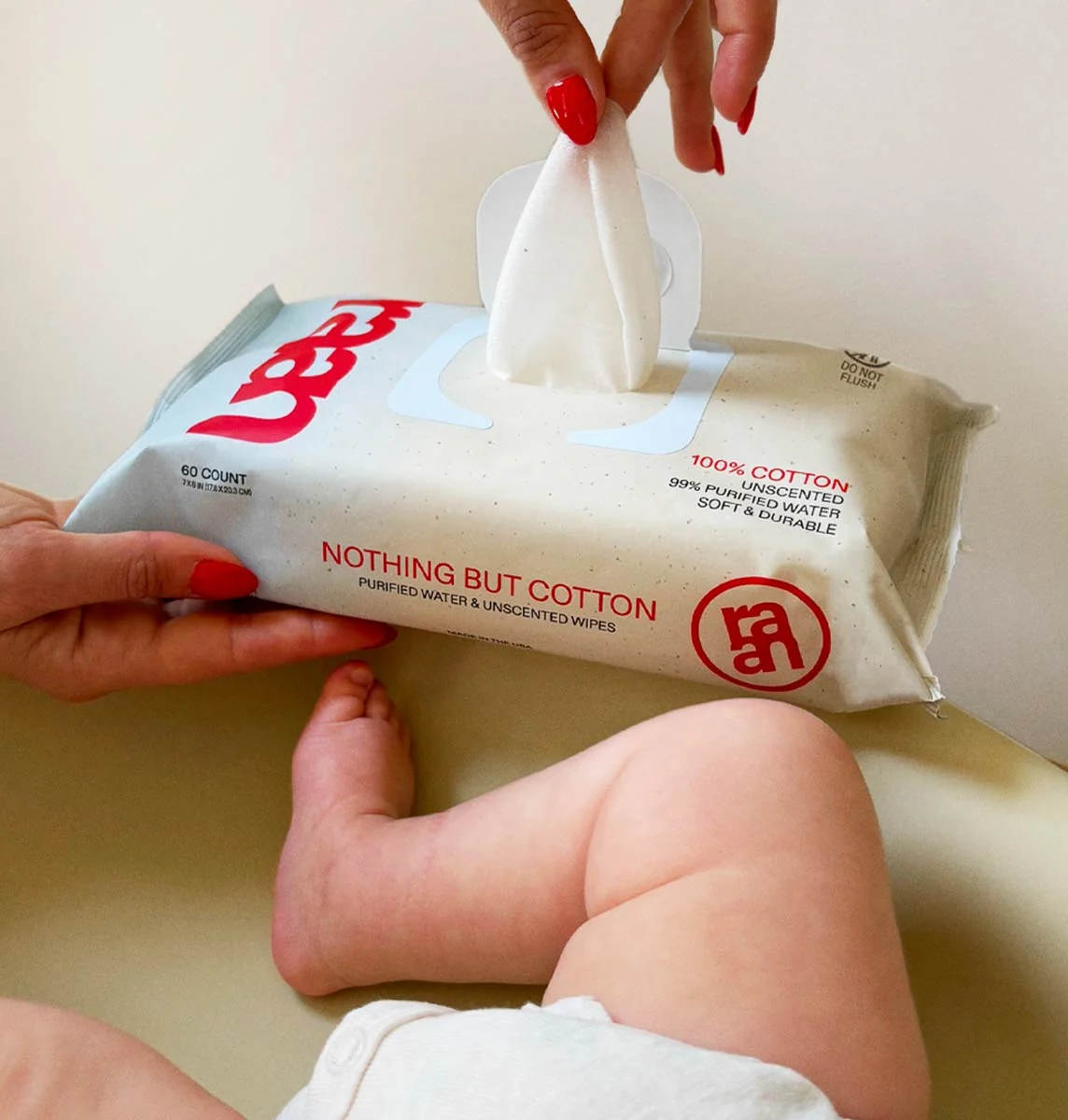 A hand with red nails pulls a wipe from a package of Nothing But Cotton wipes next to a baby.